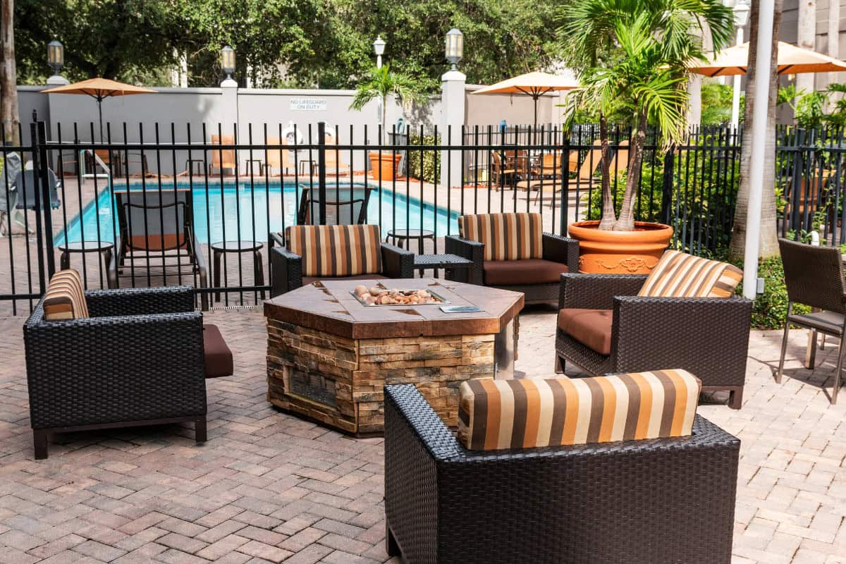 fire-pit-under-covered-patio-installation Cozy outdoor seating area with wicker chairs, striped cushions, and a fire pit near a swimming pool, surrounded by lush tropical plants and umbrellas at Backyard Toasty.